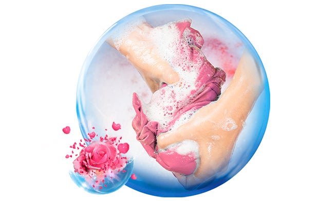 Manos enjabonando una prenda rosa con abundante espuma, resaltando una limpieza profunda y un aroma fresco a rosas.